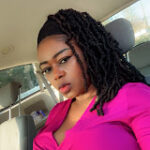 A person with long braided hair sits in a car, wearing a bright pink top and looking towards the camera.