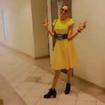 Person in a yellow dress and black boots stands in a hallway, striking a playful pose with both hands making peace signs. They are wearing sunglasses.