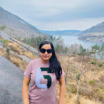A woman with sunglasses stands on a rocky hill with a scenic view of a lake and mountains in the background.