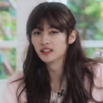 A person with long dark hair is speaking indoors, wearing a pink blazer. The background is blurred.