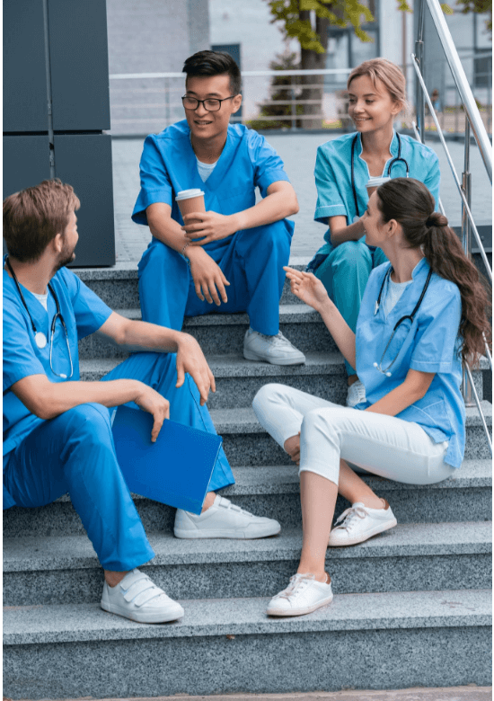 Four medical professionals in scrubs sit on outdoor steps, talking and smiling. One holds a clipboard, another holds a coffee cup.