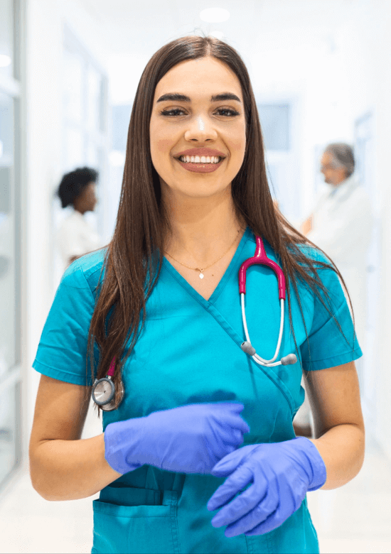 A healthcare worker in blue scrubs and purple gloves stands in a hallway, smiling, with a stethoscope around her neck. Other people are blurred in the background.
