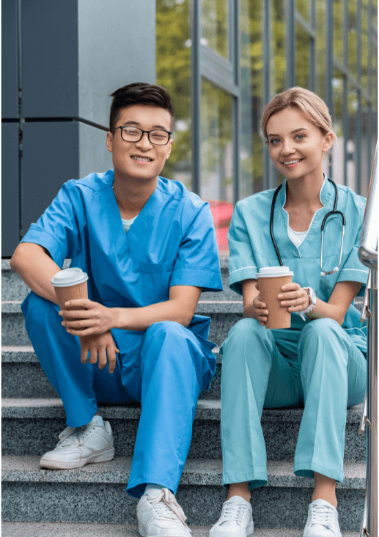 Two healthcare professionals in scrubs sit on stairs outdoors, each holding a disposable coffee cup and smiling at the camera.