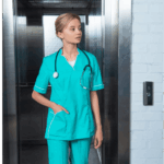 A healthcare worker in teal scrubs with a stethoscope around their neck steps out of an elevator in a medical facility.